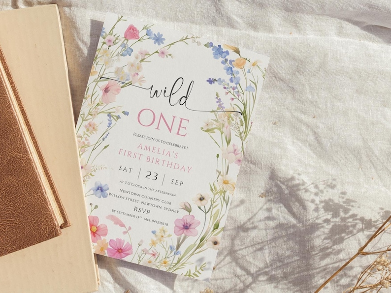 May include: A floral-themed invitation for a first birthday celebration. The card features the text "wild ONE" in a script font, with details about the event, including the date and location, surrounded by a watercolor floral border with pink, blue, and yellow flowers.