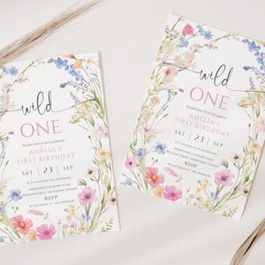 May include: Two floral-themed birthday invitations with the text "wild ONE" in a handwritten font. The invitations feature a watercolor floral border with pink, blue, and white flowers. The text includes the date, time, and location of the party.
