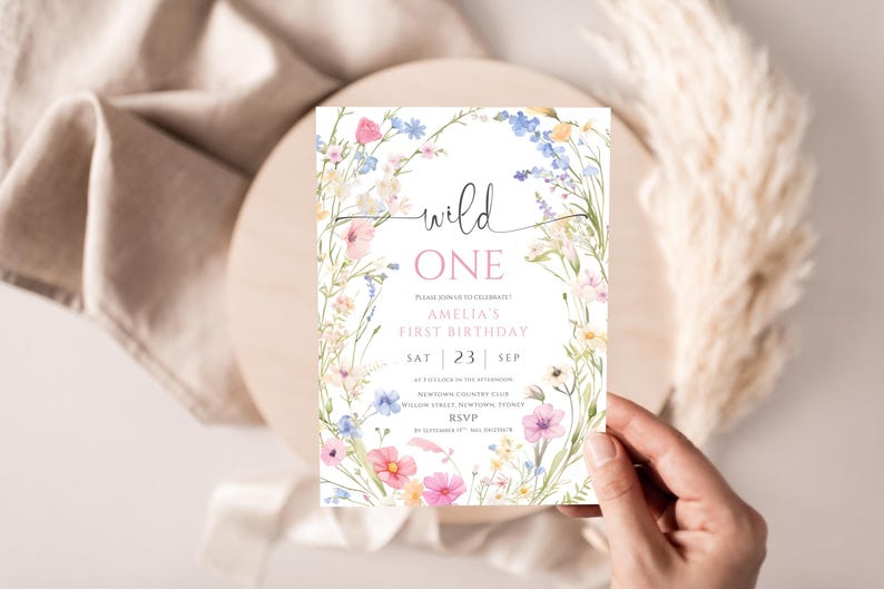 May include: A floral-themed birthday invitation with the words "wild ONE" in a script font. The invitation features a watercolor design of pink, blue, and yellow flowers. The text includes the name "Amelia", the date, and RSVP information.