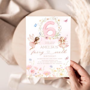 May include: A fairy-themed birthday invitation with a pink number 6 surrounded by flowers. The invitation features two fairy illustrations, butterflies, and the text "Amelia's fairy sixth birthday". The invitation is held by a hand.