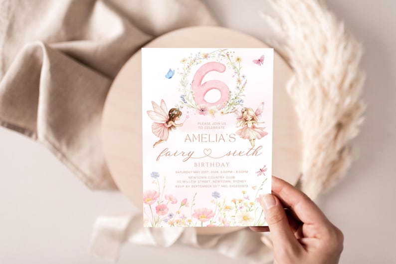 May include: A fairy-themed birthday invitation with a pink number 6 surrounded by flowers. The invitation features two fairy illustrations, butterflies, and the text "Amelia's fairy sixth birthday". The invitation is held by a hand.