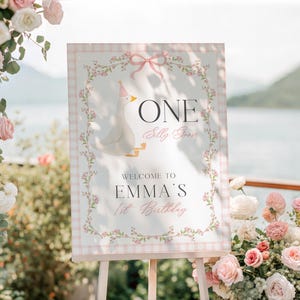May include: A decorative sign on an easel with a light pink gingham border and floral accents. The sign reads "ONE Silly Goose" and "Welcome to Emma's 1st Birthday" with a cartoon goose illustration. The background includes flowers and a scenic view.