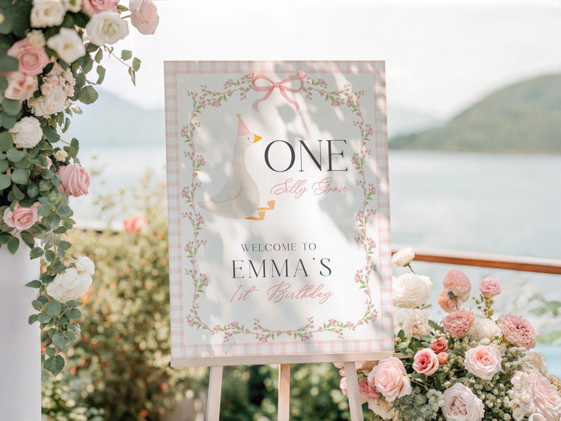 May include: A decorative sign on an easel with a light pink gingham border and floral accents. The sign reads "ONE Silly Goose" and "Welcome to Emma's 1st Birthday" with a cartoon goose illustration. The background includes flowers and a scenic view.