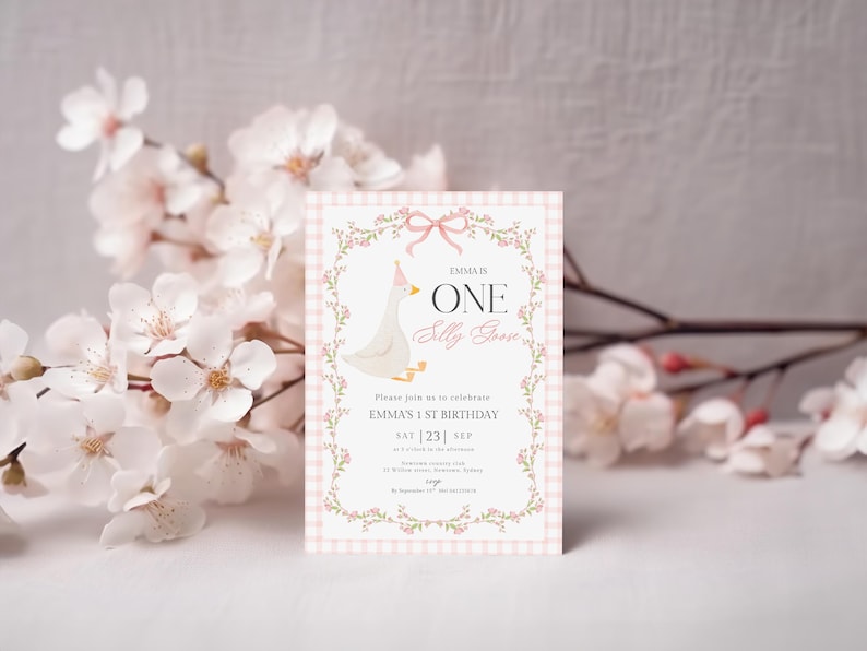 May include: A birthday invitation with a pink gingham border and floral accents. The invitation features a cartoon goose wearing a party hat and the text "Emma is One". The invitation is surrounded by white flowers.