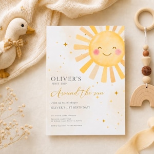 Puede incluir: Una invitación para el primer cumpleaños de un bebé con una ilustración de sol en acuarela. La invitación dice "Oliver's First Trip Around the Sun" en escritura dorada. Un juguete de pato de peluche y un móvil de bebé de madera también están en la imagen.