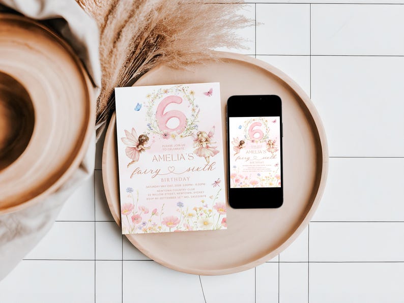 May include: A pink and floral birthday invitation with the number 6, fairies, and the text "Amelia's fairy sixth birthday" is displayed on a wooden tray next to a smartphone with the same design. Dried flowers and a wooden bowl are also in the image.