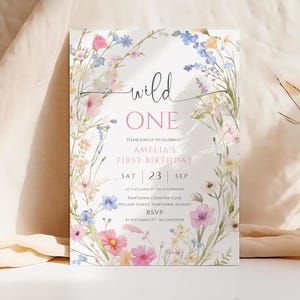 May include: A floral-themed invitation for a first birthday celebration. The text "wild ONE" is displayed in a delicate script, with the details of the event, including the date, time, and location, written below. The invitation is adorned with watercolor flowers.