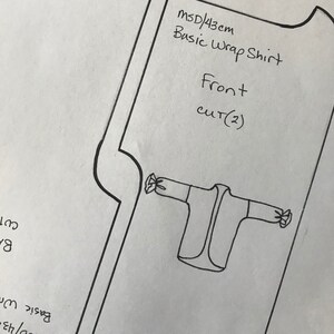 May include: A black and white line drawing of a wrap shirt pattern on a white piece of paper. The pattern is labeled "MSD/43cm Basic Wrap Shirt Front CUT(2)".