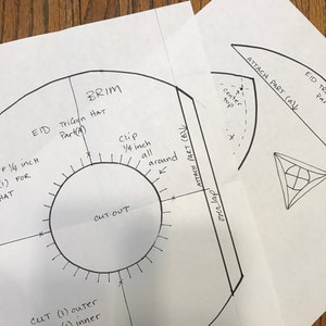 May include: White paper pattern pieces with handwritten text and diagrams for a tricorn hat. The text includes "BRIM", "CUT OUT", "Clip 1/4 inch all around", and measurements in inches. The pattern is for a hat.