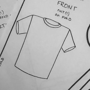 May include: A black and white line drawing of a t-shirt with a crew neck and short sleeves. The text "FRONT cut (1) on FOLD" is written above the t-shirt.