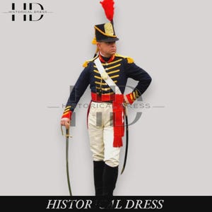 Könnte beinhalten: Eine historische Militäruniform bestehend aus einer dunkelblauen Jacke mit gelber Flechtverzierung, einer weißen Hose und einem schwarzen Tschako mit roter Feder. Das Set umfasst einen weißen Kreuzgurt, eine rote Schärpe und einen gebogenen Säbel.