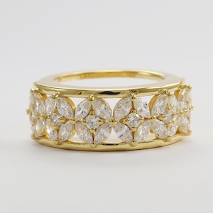 May include: A gold band ring featuring a repeating pattern of marquise-cut clear stones arranged in circular clusters. The design includes two thin horizontal gold rails at the top and bottom, framing the central row of sparkling white gemstones.