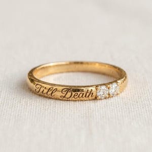 May include: A gold band ring featuring a textured surface with the engraved script text Till Death and two small round clear stones set side by side on the outer circumference of the metal band, creating a simple and elegant jewelry piece.