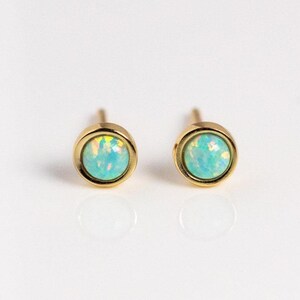 May include: A pair of stud earrings featuring round, iridescent light blue opals set in polished gold-toned metal bezels. The stones display internal flashes of green and yellow light, and each earring includes a visible metal post on the back.