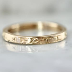 Puede incluir: Un anillo dorado con la palabra "FAT" grabada en la banda. El anillo tiene un diseño simple y elegante, con un acabado liso y pulido. El anillo probablemente está hecho de oro o metal dorado.
