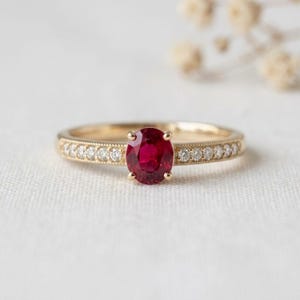 Könnte beinhalten: Ein Goldring mit einem zentralen, oval geschliffenen, tiefroten Rubin, der von vier Krappen gehalten wird. Die Ringschiene ist auf beiden Seiten mit einer Reihe kleiner, runder, klarer weißer Diamanten besetzt, die in einer feinen Milgrain-Fassung liegen.