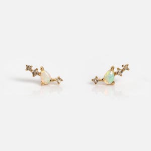 May include: A pair of gold stud earrings featuring a central pear-shaped opal stone flanked by two small clear round stones on each side. The metal setting is polished gold, and the design follows a curved, elongated shape that sits along the earlobe.