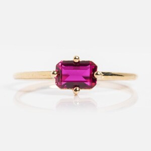 Op de afbeelding: Een gouden ring met een rechthoekige, felroze edelsteen. De edelsteen is gezet met vier kleine gouden accenten. De ring heeft een eenvoudige, gepolijste gouden band. Het ontwerp is minimalistisch en elegant.
