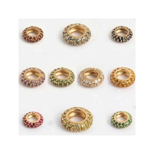 Puede incluir: Nueve anillos dorados, cada uno incrustado con piedras preciosas de colores. Los anillos presentan una variedad de colores, incluyendo rojo, verde, morado y amarillo. Las piedras preciosas están engastadas muy juntas, creando un efecto brillante. Los anillos se exhiben sobre una superficie blanca.