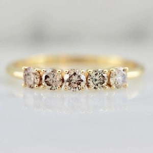Peut inclure: Une bague en or ornée de cinq diamants ronds brun clair, disposés côte à côte. Les diamants scintillent, et la bague se détache sur un fond flou. Il s'agit d'un bijou élégant.