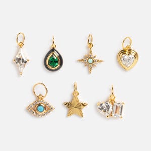 May include: A collection of gold-colored charms with various designs, including a diamond shape, a teardrop with a green stone, a starburst, a heart, an eye, a star, and a combination of heart and rectangular shapes. Each charm has a small loop for attachment.