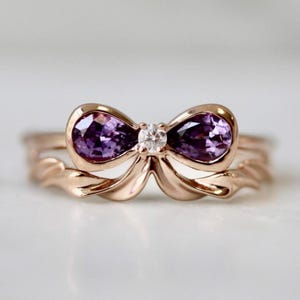 Könnte beinhalten: Ein roségoldener Ring mit einem Schleifendesign. Der Bogen ist mit zwei oval geschliffenen lila Edelsteinen und einem kleinen, runden, klaren Edelstein verziert. Zartes, feminines Design.