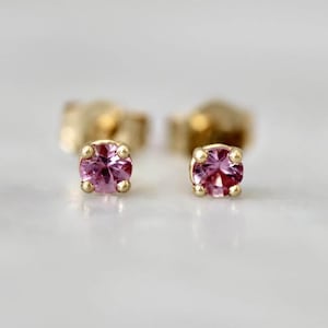 May include: A pair of stud earrings featuring round-cut pink gemstones set in four-prong yellow gold settings. The stones are faceted, reflecting light with a vibrant magenta hue, while the gold prongs securely hold each stone in place.