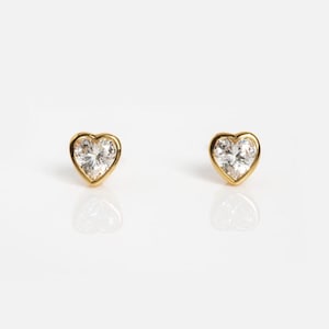 May include: A pair of stud earrings featuring heart-shaped clear stones set in gold-toned metal. Each earring displays a faceted, brilliant-cut stone framed by a smooth, polished gold border, creating a classic and symmetrical appearance.