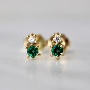 May include: A pair of gold stud earrings, each featuring a small, clear, round-cut stone positioned above a larger, vibrant green, round-cut stone. The stones are set in gold prongs, creating a vertical, two-stone design for each earring.