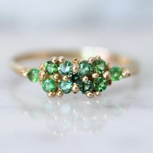 May include: A gold ring featuring a cluster of small, round, green gemstones. The gemstones are a mix of light and dark green, set in a delicate gold band. The ring has a simple, elegant design.