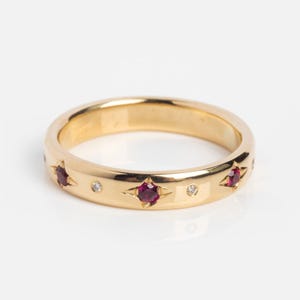 Può includere: Un anello a fascia in oro con pietre preziose a forma di stella color rubino e piccoli accenti di diamanti. L'anello ha una finitura lucida ed è posto su uno sfondo bianco. È un gioiello.