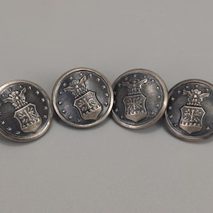 Puede incluir: Cuatro botones plateados con un emblema detallado que presenta un águila y un escudo. Los botones tienen forma circular con un fondo texturizado y están dispuestos en fila sobre un fondo gris. El emblema está en el centro de cada botón.