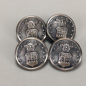 Puede incluir: Cuatro botones plateados vintage con un emblema detallado que presenta un águila, un escudo y estrellas. Los botones son redondos y tienen un diseño ligeramente en relieve, perfectos para añadir un toque de historia a la ropa o a las manualidades.