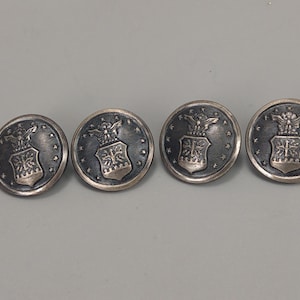 Puede incluir: Cuatro botones plateados con un diseño detallado. Cada botón presenta un emblema central con un águila y un escudo, rodeado de estrellas. Los botones tienen una apariencia ligeramente envejecida, con un diámetro de unos 2.5 cm.