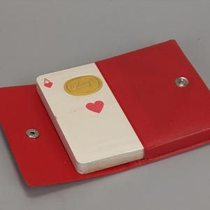 May include: A red leather playing card case with a snap closure, open to reveal a deck of cards. The cards are white with red heart symbols and the Ace of Hearts visible. The case is on a grey background.