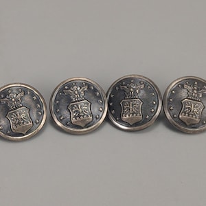 Puede incluir: Cuatro botones plateados antiguos con un emblema detallado que presenta un águila y un escudo. Los botones tienen forma circular con un diseño en relieve y una apariencia oscura y envejecida, lo que sugiere que son vintage. Cada botón mide aproximadamente 2,5 cm de diámetro.