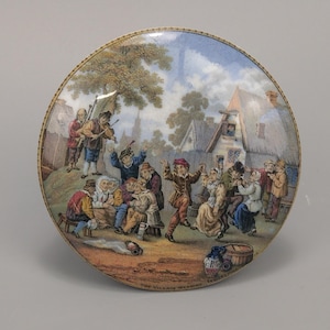 Puede incluir: Un plato decorativo redondo con una ilustración detallada y colorida de una escena de pueblo. La imagen representa a personas bailando y tocando música, con edificios y árboles al fondo. El plato tiene un borde dorado y el texto "The Village Wedding" en la parte inferior.