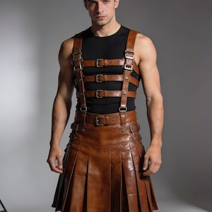 May include: A brown leather kilt with pleats and multiple buckles, paired with a matching leather harness. The harness features straps and buckles across the chest and shoulders. The outfit is designed with a rugged, yet stylish aesthetic.