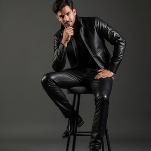 May include: A black leather jacket and matching pants. The jacket has a classic design with a collar and zipper. The pants are form-fitting. The person is wearing black boots and is seated on a black stool against a gray background.