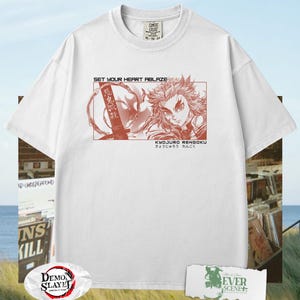 May include: White t-shirt featuring a red graphic of a character from the anime series Demon Slayer. The graphic includes the text "SET YOUR HEART ABLAZE" and Japanese text. The shirt has a crew neck and short sleeves.