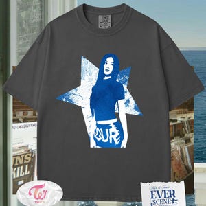 May include: Dark grey t-shirt with a blue graphic of a woman inside a star. The word "TOUR" is visible on the bottom of the graphic. The shirt has a crew neck and short sleeves. The brand name "Comfort Colors" is printed on the shirt.