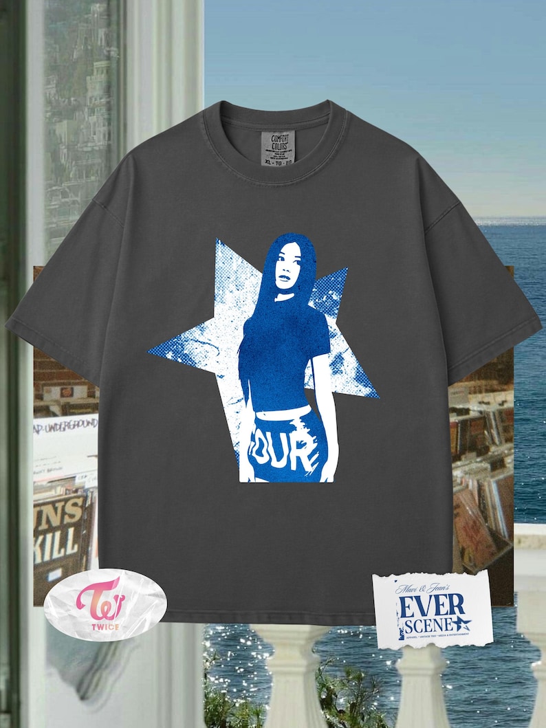 May include: Dark grey t-shirt with a blue graphic of a woman inside a star. The word "TOUR" is visible on the bottom of the graphic. The shirt has a crew neck and short sleeves. The brand name "Comfort Colors" is printed on the shirt.