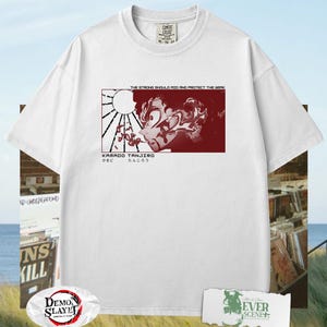 May include: White t-shirt featuring a red graphic of an anime character with Japanese text. The graphic includes the text "THE STRONG SHOULD RID AND PROTECT THE WEAK" and "KAMADO TANJIRO". The shirt has a crew neck and short sleeves.
