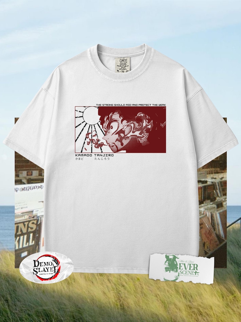 May include: White t-shirt featuring a red graphic of an anime character with Japanese text. The graphic includes the text "THE STRONG SHOULD RID AND PROTECT THE WEAK" and "KAMADO TANJIRO". The shirt has a crew neck and short sleeves.