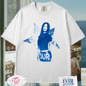 May include: White t-shirt featuring a blue graphic of a woman and the word "TOUR". The design is set against a starburst background. The shirt has a crew neck and short sleeves. The tag on the shirt reads "COMFORT COLORS".