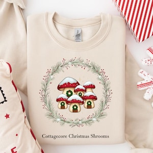 May include: A beige long-sleeve sweatshirt featuring a graphic of five mushrooms with red caps and white spots, arranged inside a green pine wreath. The text "Cottagecore Christmas Shrooms" is printed in black below the wreath design.