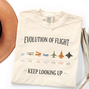 May include: A cream-colored t-shirt featuring the text "EVOLUTION OF FLIGHT" and "KEEP LOOKING UP" in black. Between the text is a timeline graphic showing various aircraft including a paper airplane, biplane, jetliner, and a future concept design.