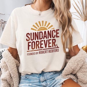 May include: Cream-colored t-shirt with the text "SUNDANCE FOREVER" in burgundy and a sunburst graphic. The text "FOUNDED BY ROBERT REDFORD" is below the main text. The shirt is short-sleeved.