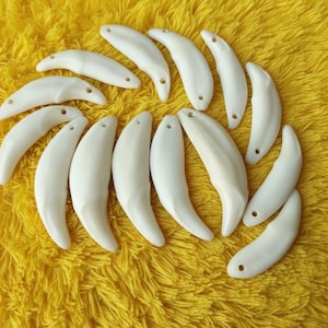 Könnte beinhalten: Eine Sammlung von cremeweißen, gebogenen Anhängern, die Zähnen ähneln, jeweils mit einem kleinen Loch an einem Ende. Die Anhänger sind auf einer leuchtend gelben, strukturierten Oberfläche angeordnet. Die Anhänger haben ein glattes, poliertes Aussehen.