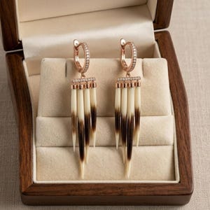 Puede incluir: Un par de pendientes de aro en oro rosa con una hilera de peque&ntilde;as piedras transparentes y cuatro p&uacute;as colgantes puntiagudas de color crema con puntas de color marr&oacute;n oscuro. Los pendientes se presentan en un joyero de madera con forro de terciopelo.